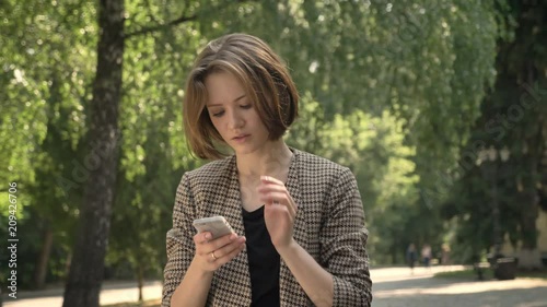 Young attractive concentrated girl is typing message on smartphone in park in daytime, in summer, communication concept photo