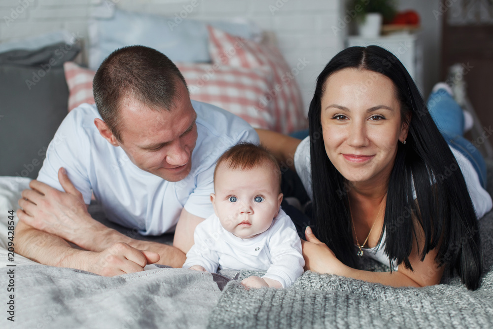 Obraz premium Young mom and dad and their baby son lie in bed and look at the camera indoors. The concept of a happy family