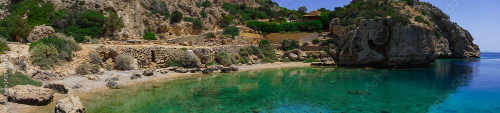 Panorama of the L- shaped stoa and a beautiful small beach beneath at archaeological site of Heraion, sanctuary of goddess Hera, in Perachora, Loutraki, Greece Panorama of the L- shaped stoa and a beautiful small beach beneath at archaeological site of Heraion, sanctuary of goddess Hera, in Perachora, Loutraki, Greece
