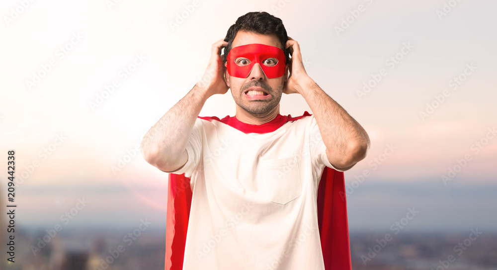 Naklejka premium Superhero man with mask and red cape takes hands on head because has migraine on a sunset background
