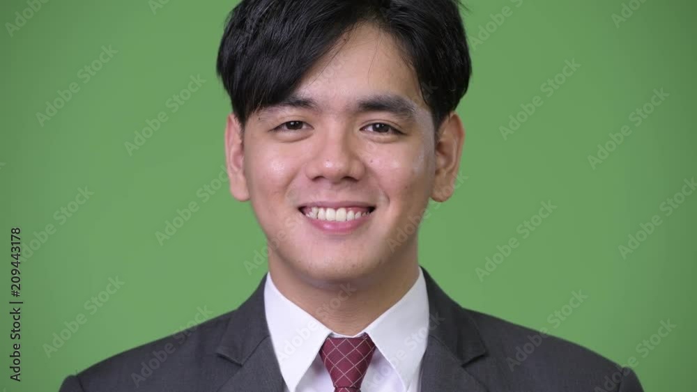 Happy young handsome Asian businessman smiling against green background Happy young handsome Asian businessman smiling against green background