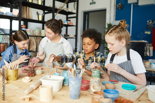 Group of creative kids and their teacher painting handmade clay items at lesson of handcraft
