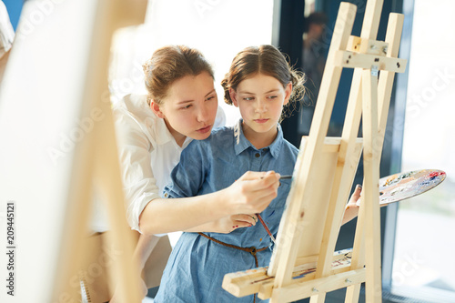 Young teacher showing one of pupils what should be corrected in her painting while pointing at easel