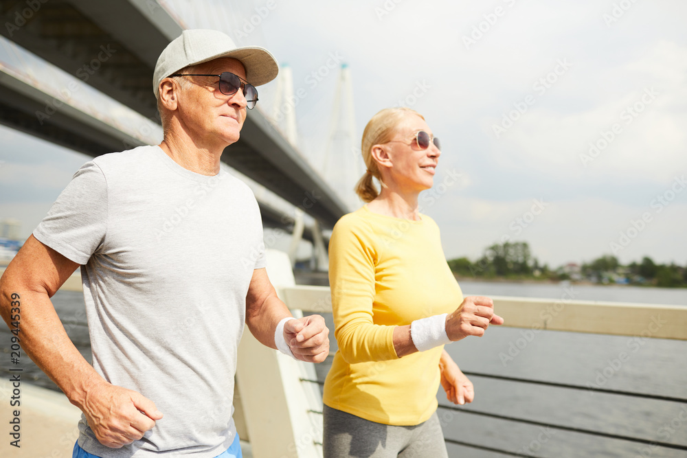 Mature spouses in activewear jogging together on sunny morning in urban environment
