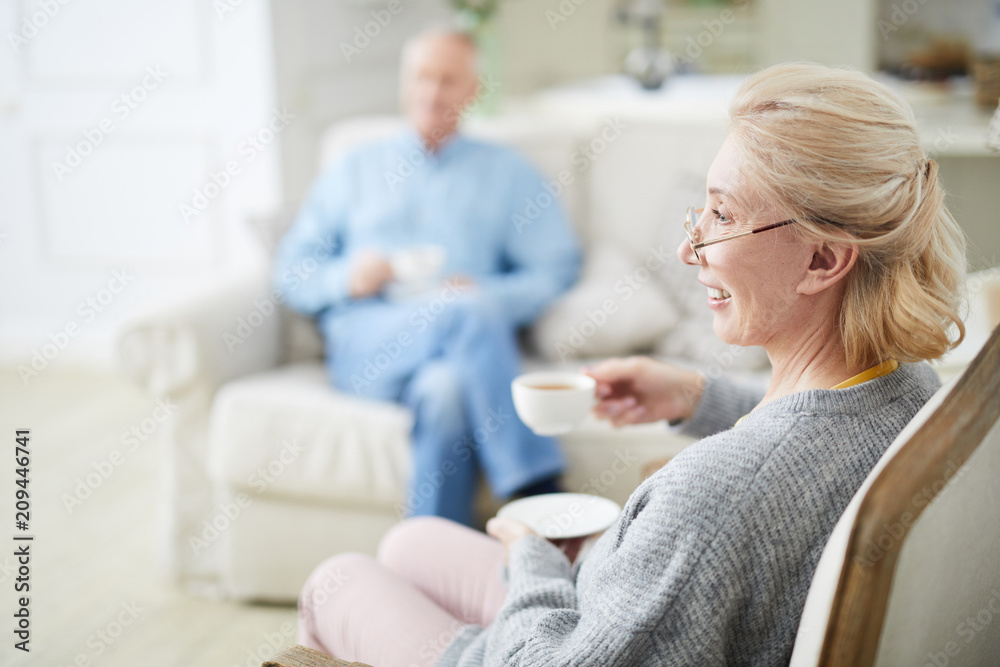 © pressmaster - Happy senior female with cup of tea sitting in armchair and watching television with her husband on background