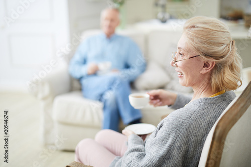 Happy senior female with cup of tea sitting in armchair and watching television with her husband on background