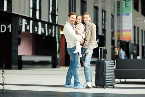 Happy couple with daughter standing inside modern airport with large suitcase near by