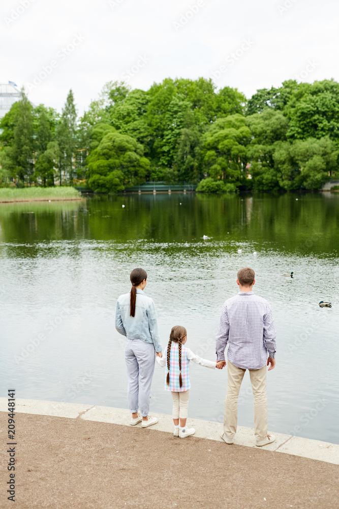 Little girl holding her parents by hands while family standing by waterside in modern park Little girl holding her parents by hands while family standing by waterside in modern park