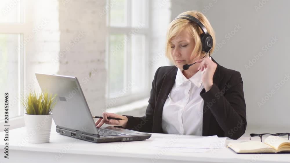 Blonde woman dressed in black jacket and white shirt working as customer call center operator, help line on laptop via headset in white office Blonde woman dressed in black jacket and white shirt working as customer call center operator, help line on laptop via headset in white office