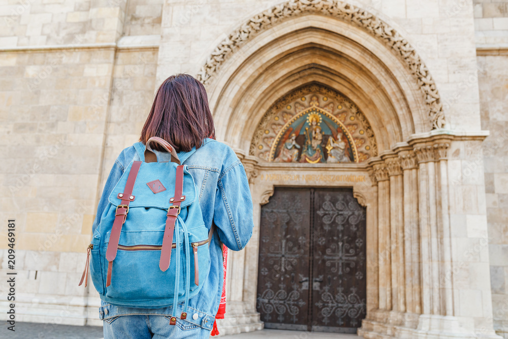 rear view of a tourist woman with backpack looking at historical architecture of a catholic gothic church, sightseeing trip concept