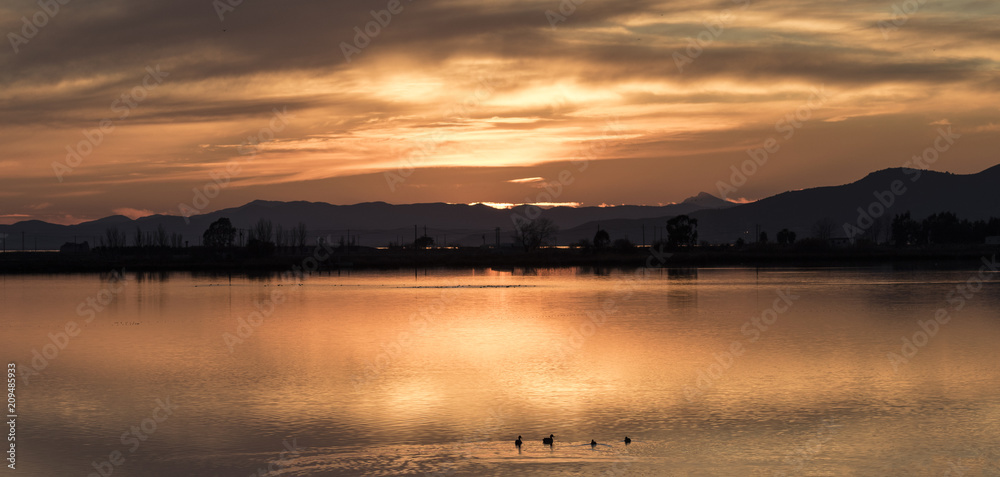 Rojiza puesta de sol entre nubes, montaña y agua de una laguna con patos nadando en la orilla, Parque natural del Delta del Ebro en Tarragona, Cataluña.