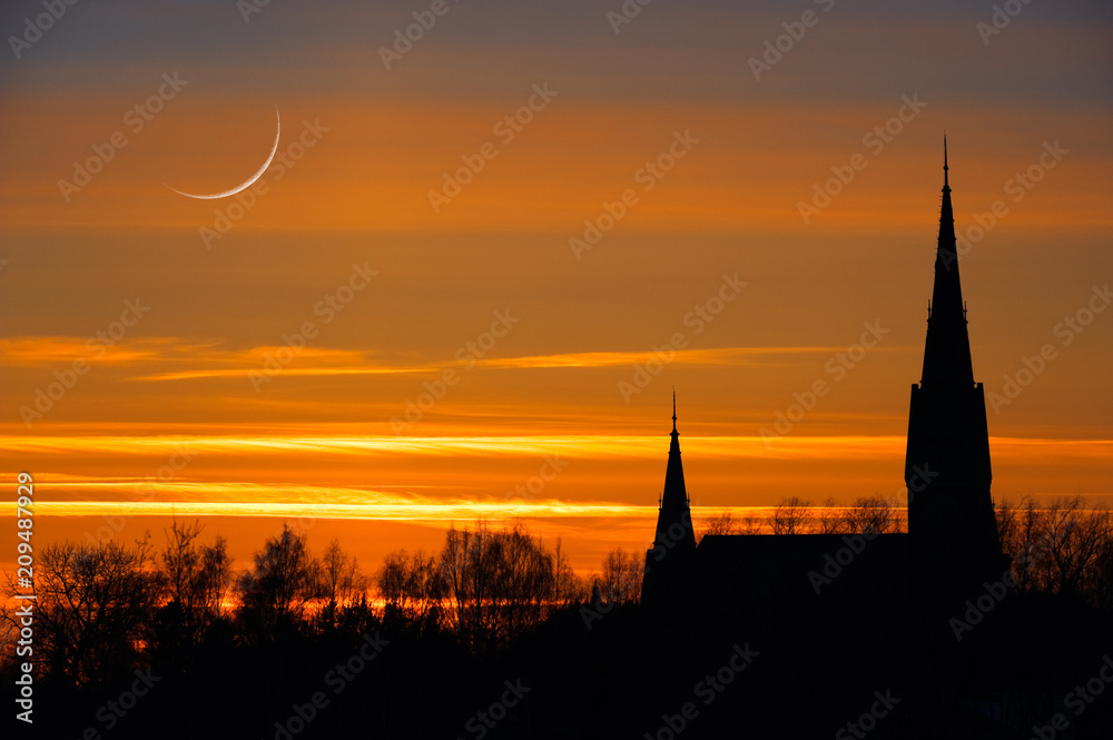 Crescent moon in twilight sky. Sunset clouds behind church towers and treetop silhouettes.