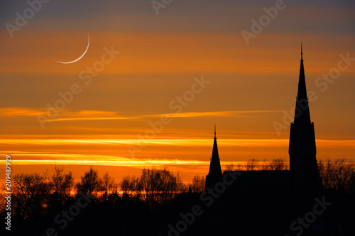 Crescent moon in twilight sky. Sunset clouds behind church towers and treetop silhouettes.