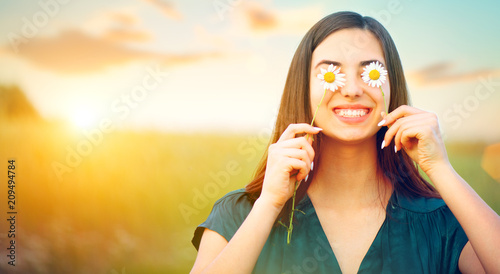 Beauty joyful girl with daisy flowers on her eyes enjoying nature and laughing on summer field. Beautiful young woman having fun