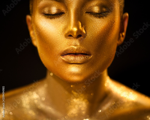 Golden skin woman face. Fashion art portrait closeup. Model girl with holiday golden glamour shiny professional makeup. Gold jewellery, jewelry, accessories. Beauty gold metallic body, lips and skin