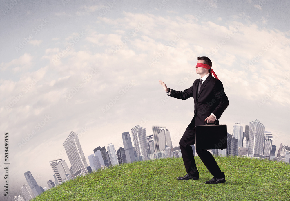 Young blindfolded businessman steps on a a patch of grass with a grey buildings in the background Young blindfolded businessman steps on a a patch of grass with a grey buildings in the background