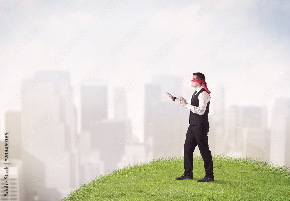 Young blindfolded businessman steps on a a patch of grass with a city in the background Young blindfolded businessman steps on a a patch of grass with a city in the background