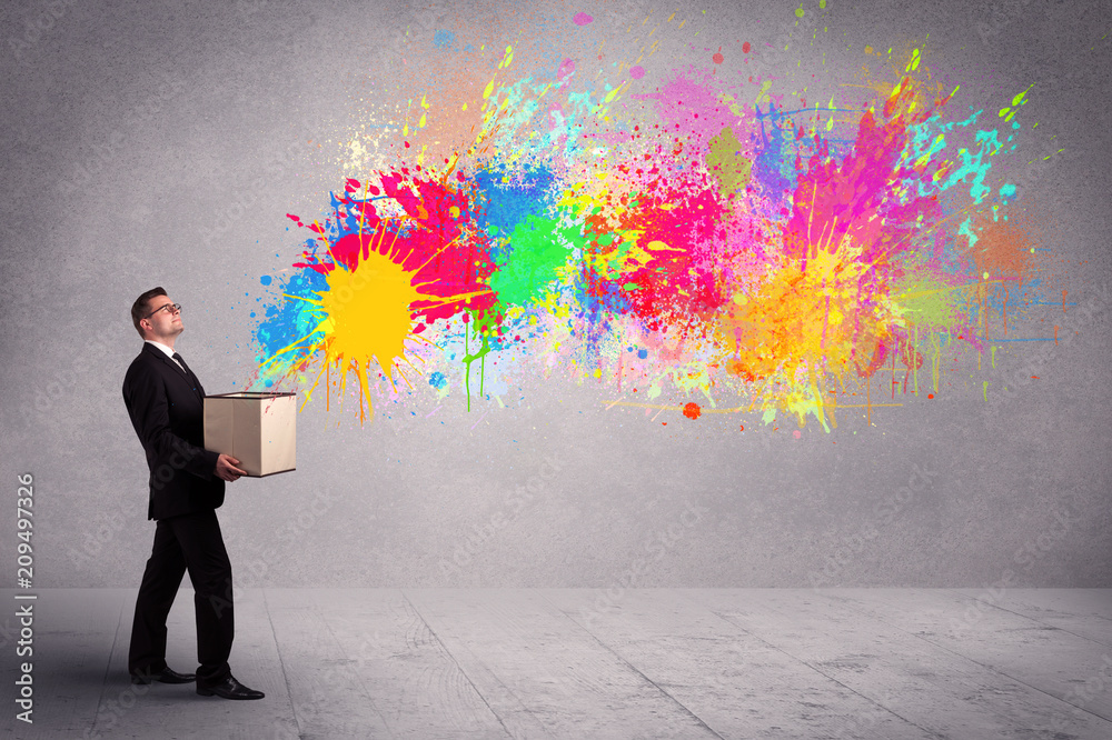 A young smiling business male holding a paperboard box with illustration of colourful spray paint splash on urban wall background concept. A young smiling business male holding a paperboard box with illustration of colourful spray paint splash on urban wall background concept.
