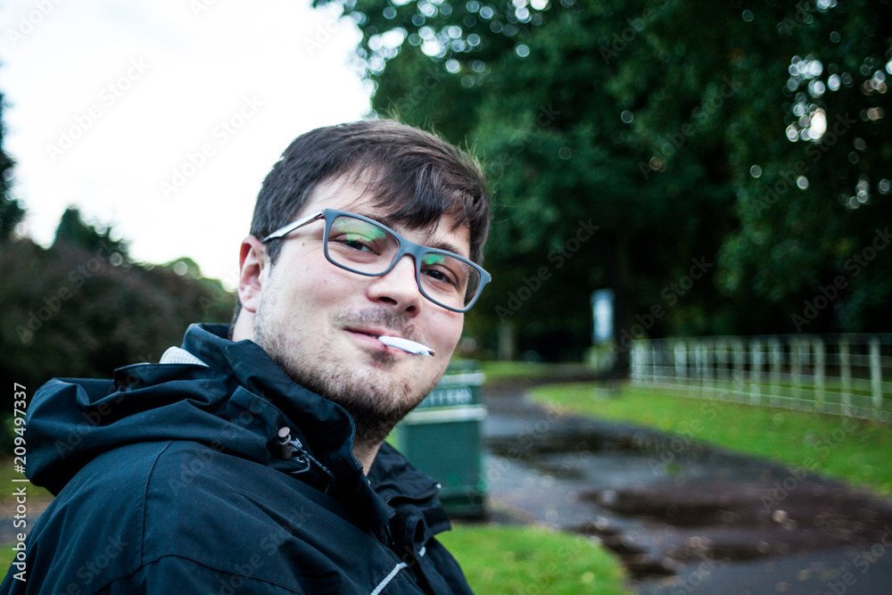 Young man with glasses and cigarette in his mouth taking selfie image with blurred background. Happy man smiling into camera and smoking a cigar. Taking selfie images in the nature.