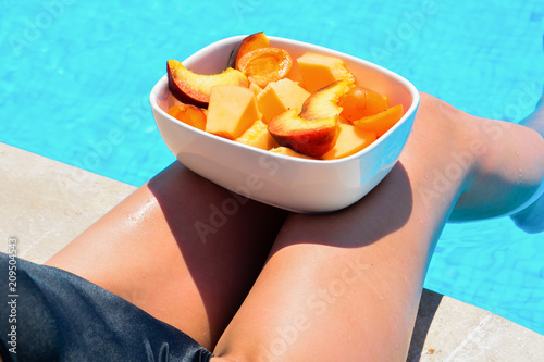 Tanned beautiful legs. Young woman relaxing and eating fruits. Fruits in a bowl by the pool near a thin slender fit girl legs. Fruit salad, healthy food. Summer concept.
