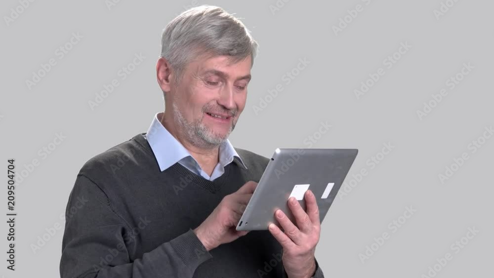 Smiling caucasian man using digital tablet. Cheerful mature man talking via internet on grey background. People, technology, communication. Smiling caucasian man using digital tablet. Cheerful mature man talking via internet on grey background. People, technology, communication.