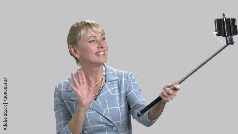 Woman holding monopod and waving with hand. Pretty smiling woman using selfie stick with a phone on a gray background. People and modern technology. Woman holding monopod and waving with hand. Pretty smiling woman using selfie stick with a phone on a gray background. People and modern technology.