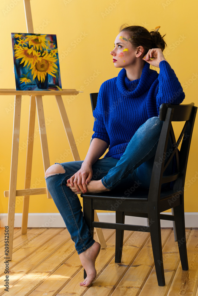 Girl artist sitting on chair in sweater and jeans. She is barefoot and with  dirty face in paint. In background picture with sunflowers on easel.  Creative profession. Stock Photo | Adobe Stock