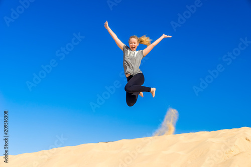 ein junges Mädchen springt vor blauem Himmel im Spagat von einer Sanddüne auf Fuerteventura