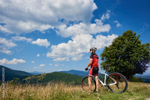 Rear view of professional bicyclist standing with bike on grassy hill. Athletic tourist enjoying beautiful view of distant mountains on summer sunny day. Healthy lifestyle and extreme sport concept.