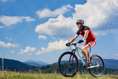 Athletic sportsman bicyclist in sportswear and helmet riding cross country bike in the Carpathian mountains, summer blue sky with clouds on background. Active lifestyle and extreme sport concept.