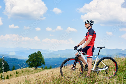 Athletic sportsman biker standing with cross country bicycle on grassy valley on summer day  enjoying beautiful view of Carpathian mountains on background. Healthy lifestyle and outdoor sport concept