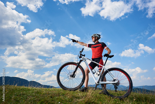 Professional male cyclist in sportswear and helmet standing with cross country bike on top of hill, pointing to the blue sky with clouds on summer sunny day. Active lifestyle and outdoor sport concept