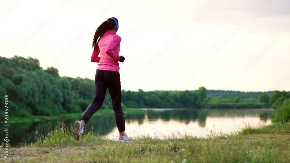 Brunette with long hair in headphones runs along the river in the Park in the morning at sunrise in the summer in a pink jacket and black pants Brunette with long hair in headphones runs along the river in the Park in the morning at sunrise in the summer in a pink jacket and black pants