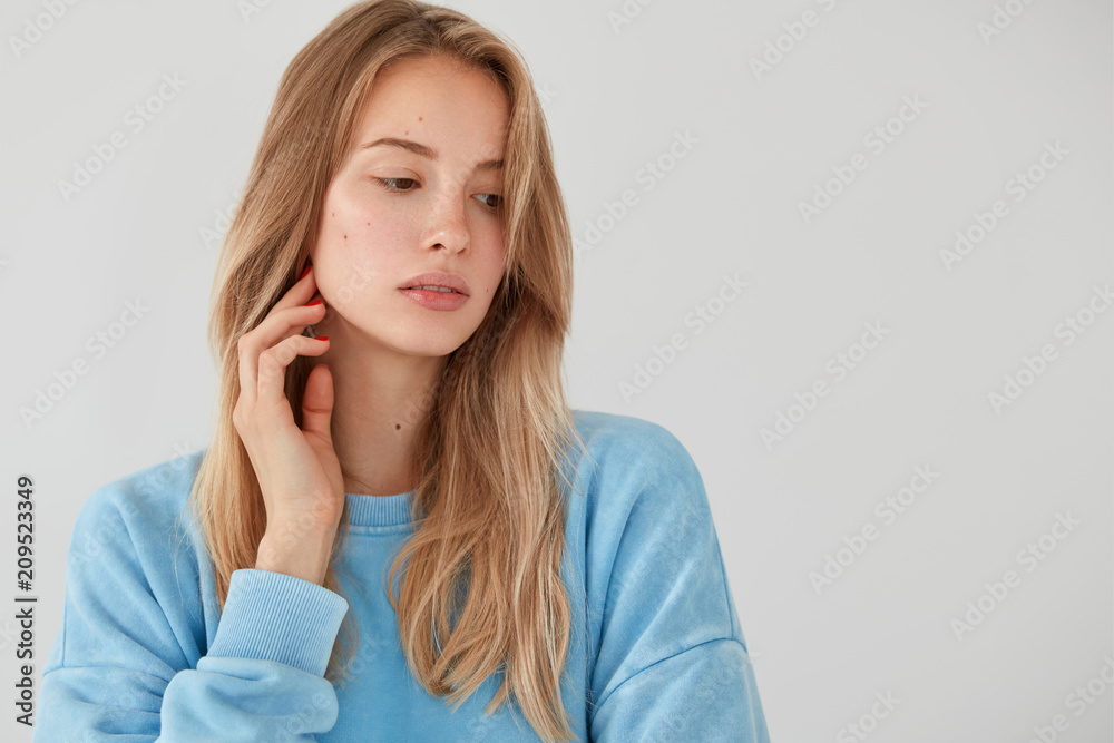 Thoughtful light haired female looks pensively aside, focused down, wears blue sweater, thinks about something, contemplates about future plans, stands against white background with blank space Thoughtful light haired female looks pensively aside, focused down, wears blue sweater, thinks about something, contemplates about future plans, stands against white background with blank space