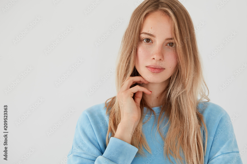 Photo of pretty female has serious look at camera, long hair, dressed in blue sweater, has gentle smile, shows her natural beauty, poses against white background with copy space for advertisement Photo of pretty female has serious look at camera, long hair, dressed in blue sweater, has gentle smile, shows her natural beauty, poses against white background with copy space for advertisement