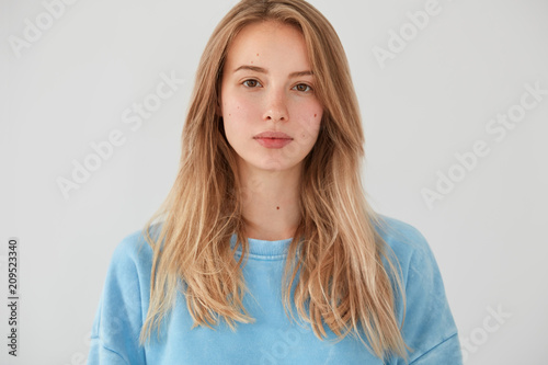 Portrait of beautiful young female with pleasant appearance looks seriously at camera, has pure skin, listens information from interlocutor, wears blue sweater, isolated over white background.