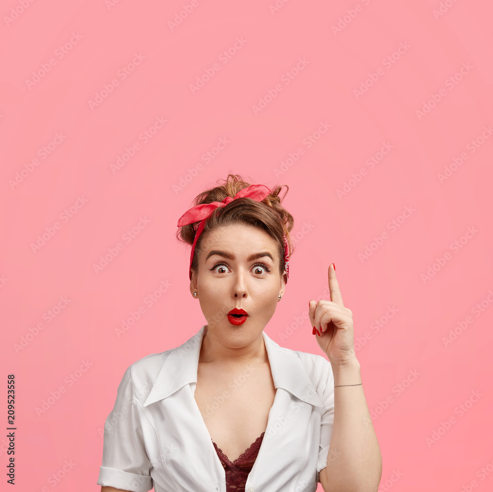 Obraz premium Vertical shot of pleasant looking stupefied female in retro style, sees something amazing, indicates with fore finger, isolated over pink background. People, advertisement and surprisement concept