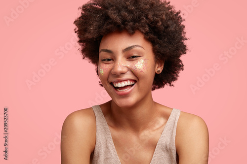 Funny happy female with Afro hairstyle  has broad smile  shows white perfect teeth  dressed in casual outfit  has sparkles on cheeks  isolated on pink background. Positive young African American woman