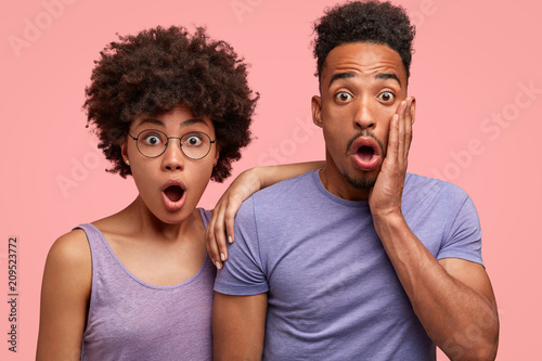 Surprised African American couple stare at camera with jaws dropped, notice or hear something unbelievable, dressed in casual t shirts, isolated over pink background. People and emotions concept