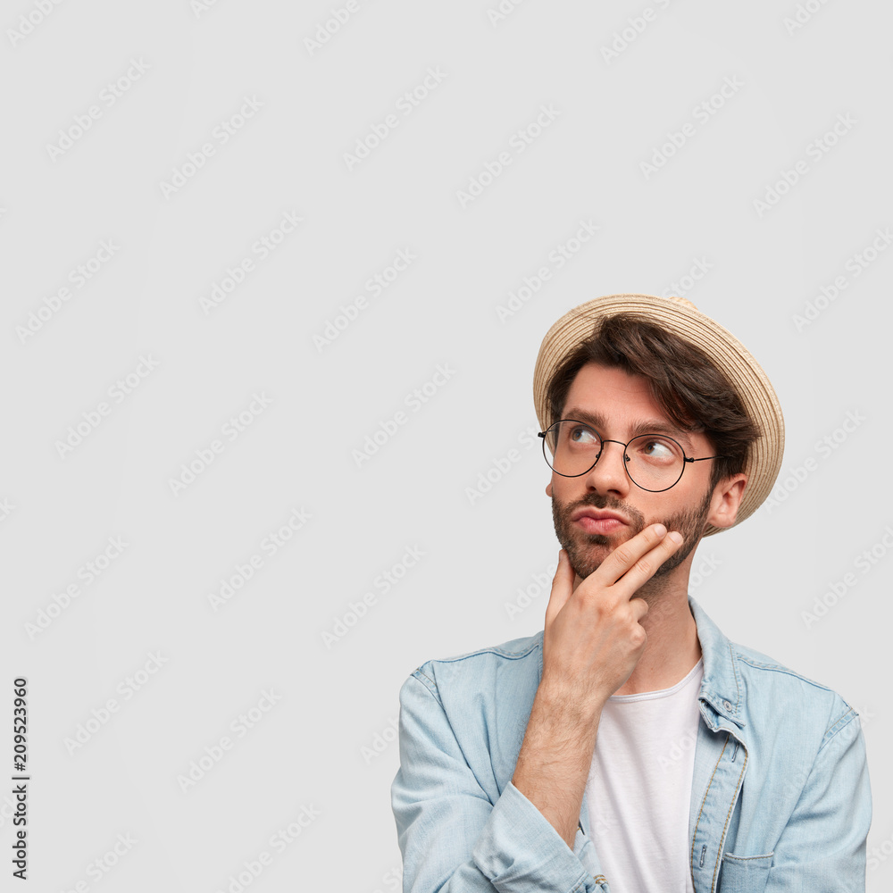 Handsome young male model holds chin, contemplates about something while looks aside, dressed in denim shirt and hat, poses against white background with copy space for your advertisement, dreams Handsome young male model holds chin, contemplates about something while looks aside, dressed in denim shirt and hat, poses against white background with copy space for your advertisement, dreams