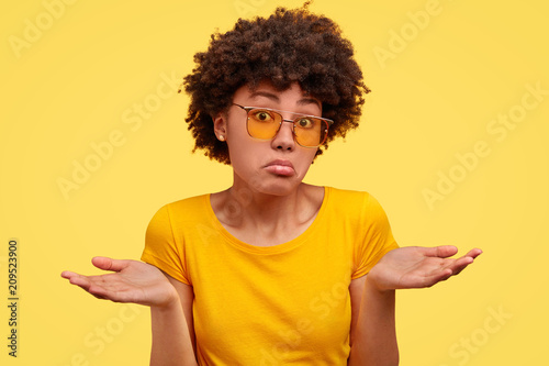 Puzzled clueless cute African American female recieves strange offer, shrugs shoulders with hesitation, feels doubt, wears bright yellow t shirt and sunglasses. People, embarrassment concept