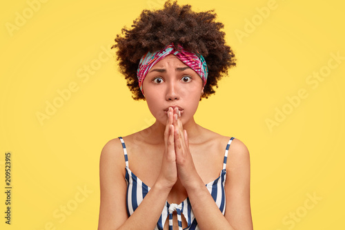 Candid shot of embarrassed cute young African American female has miserable expression, begs for something, keeps hands in praying gesture, wears striped t shirt, stands over yellow studio wall