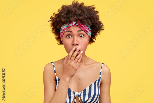 Young attractive African American female with scared expression, covers mouth and looks in puzzlement, can`t believe in latest awful news about friend, wears headband and striped casual t shirt