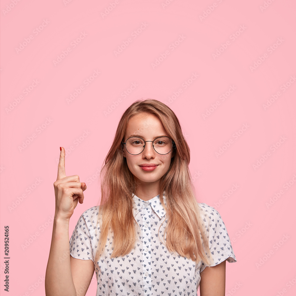 Pleased lovely young female with light hair, healthy skin, wears spectacles, indicates with fore finger upstairs, shows blank space, wears elegant blouse, being impressed by something amazing Pleased lovely young female with light hair, healthy skin, wears spectacles, indicates with fore finger upstairs, shows blank space, wears elegant blouse, being impressed by something amazing