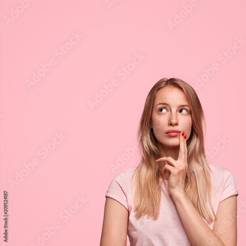 Vertical shot of thoughtful European female looks upwards, keeps fingers on cheek, contemplates about future wedding with boyfriend, dressed casually, has weekend, isolated on pink wall. Monochrome