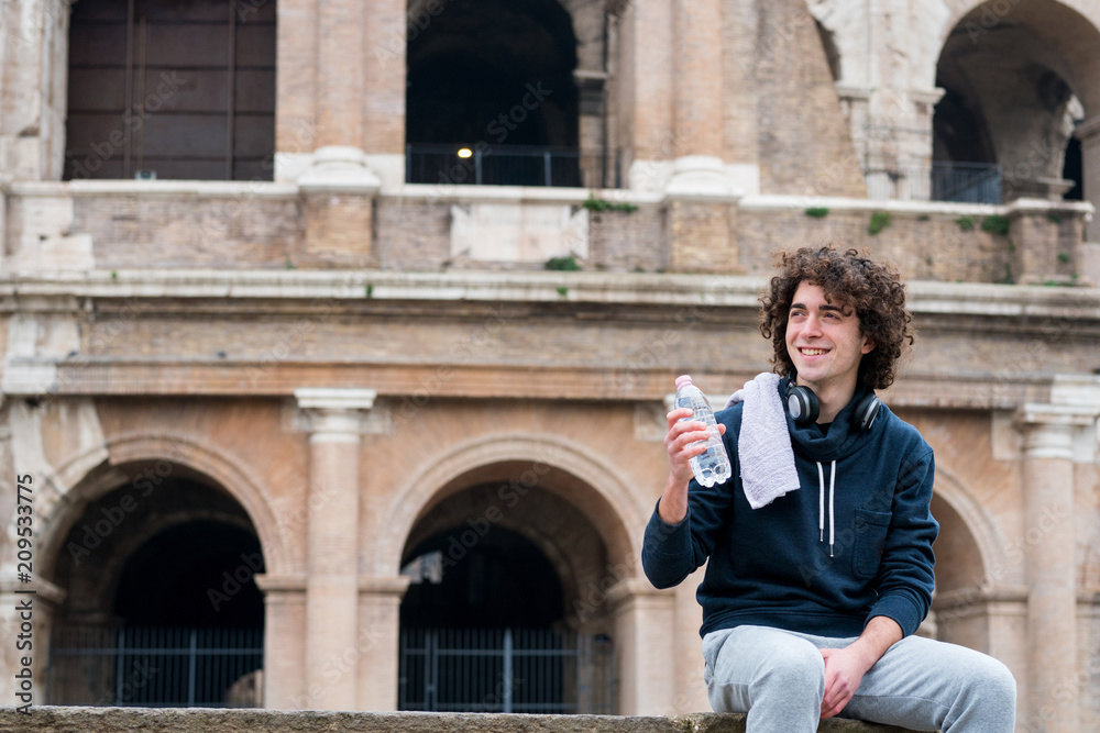 Handsome young sportsman holding water bottle and relaxing after training with a towel around his neck in front of Colosseum in Rome