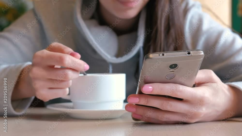 Woman is drinking a hot chocolate with spoon and look on the screen of mobile phone sitting in cafe. Closeup woman's hands. Woman is drinking a hot chocolate with spoon and look on the screen of mobile phone sitting in cafe. Closeup woman's hands.
