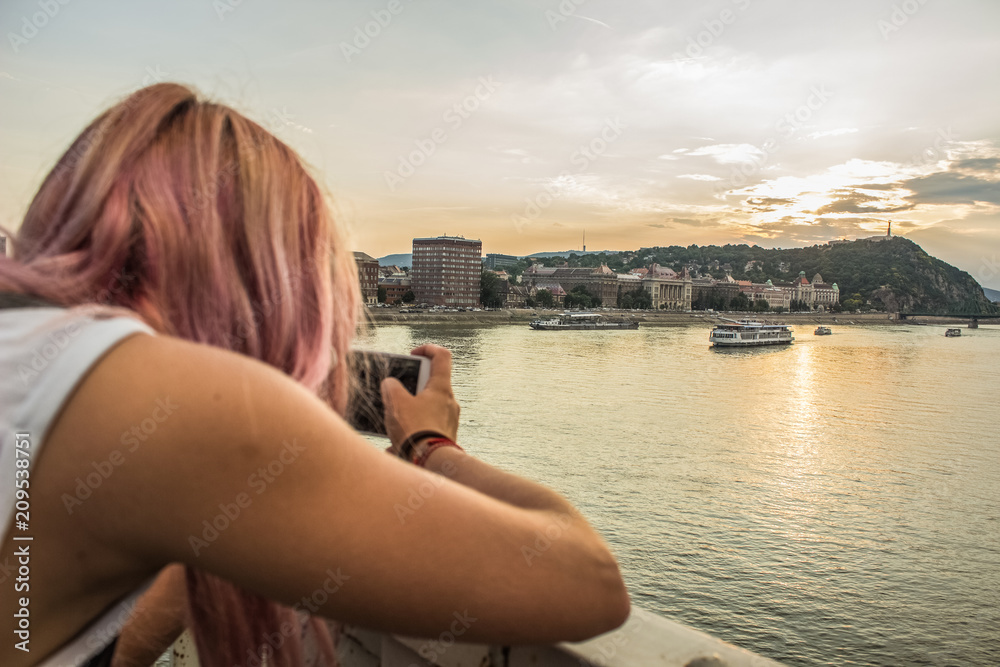 Young girl with pink hair back to camera take photo phone from Budapest waterfront urban city district in evening sunset time 