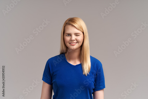 A pretty blonde girl smiles friendly and winks with a friendly smile. Studio horizontal portrait. The concept of positive emotions and feelings