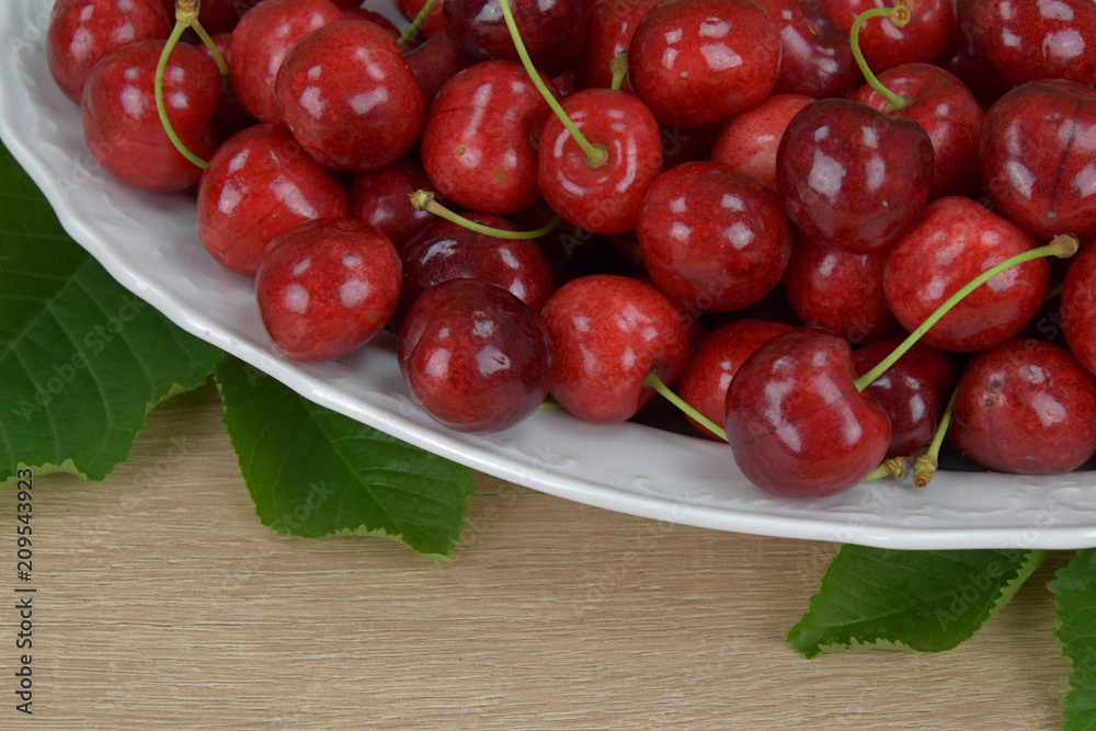 Ripe red cherries on a white plate on a wooden table. Around the green leaves. Ripe red cherries on a white plate on a wooden table. Around the green leaves.